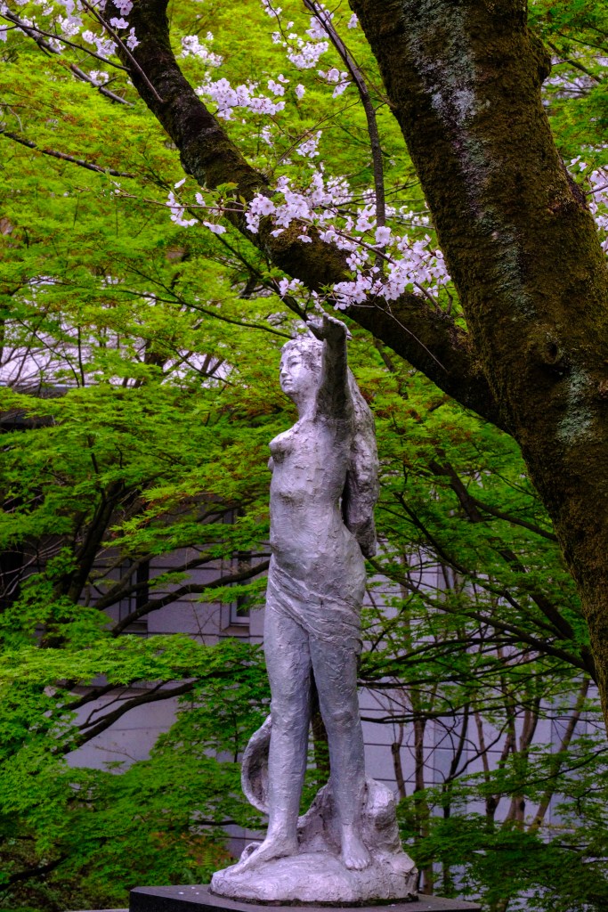 a statue in the park of a muse who seems to want to touch the sakura with one arm